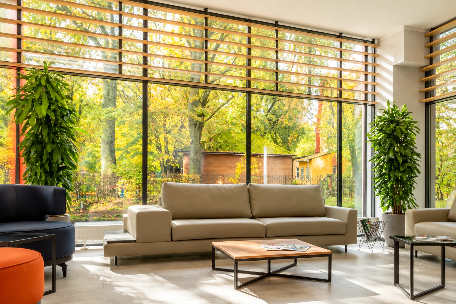 Bright four-season sunroom with beige sofas and floor-to-ceiling windows overlooking autumn trees.