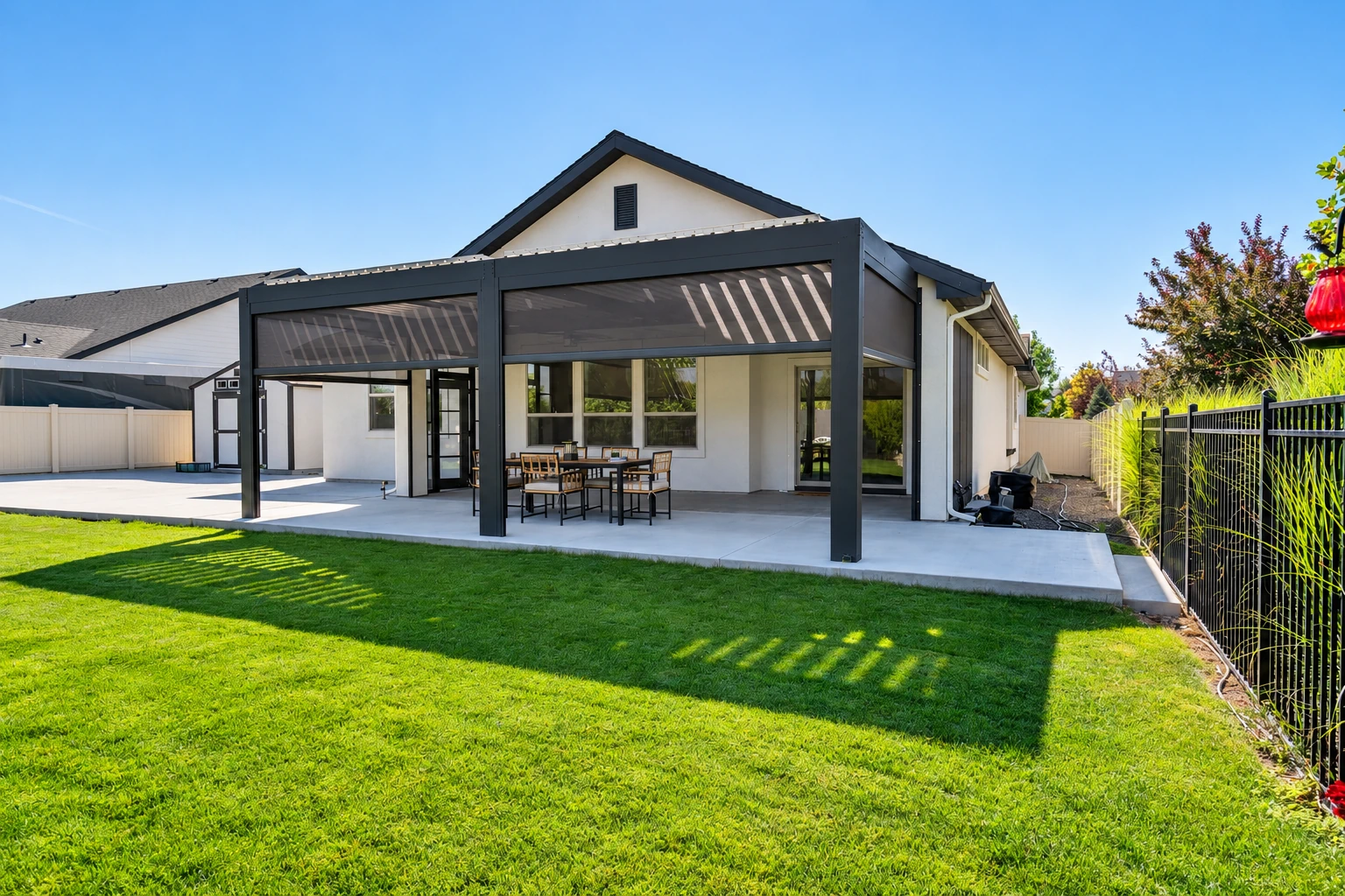 A dark-framed patio cover with mesh panels shelters an outdoor dining table beside a modern house and lawn.