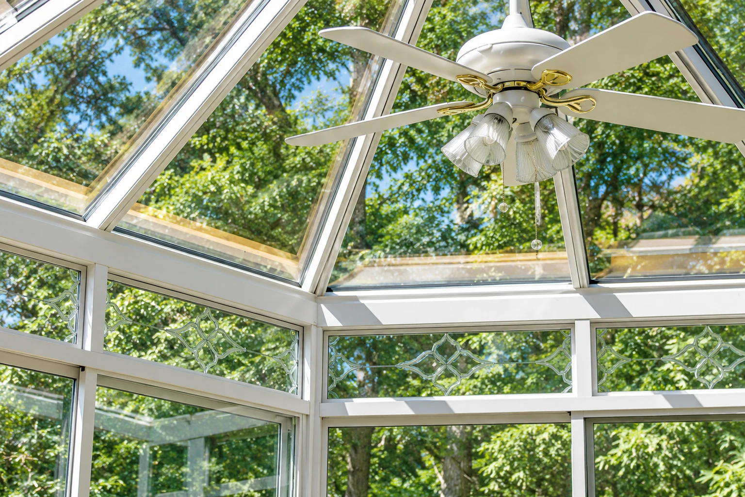 Bright four-season sunroom with a glass roof, white framing, and a ceiling fan beneath leafy trees.