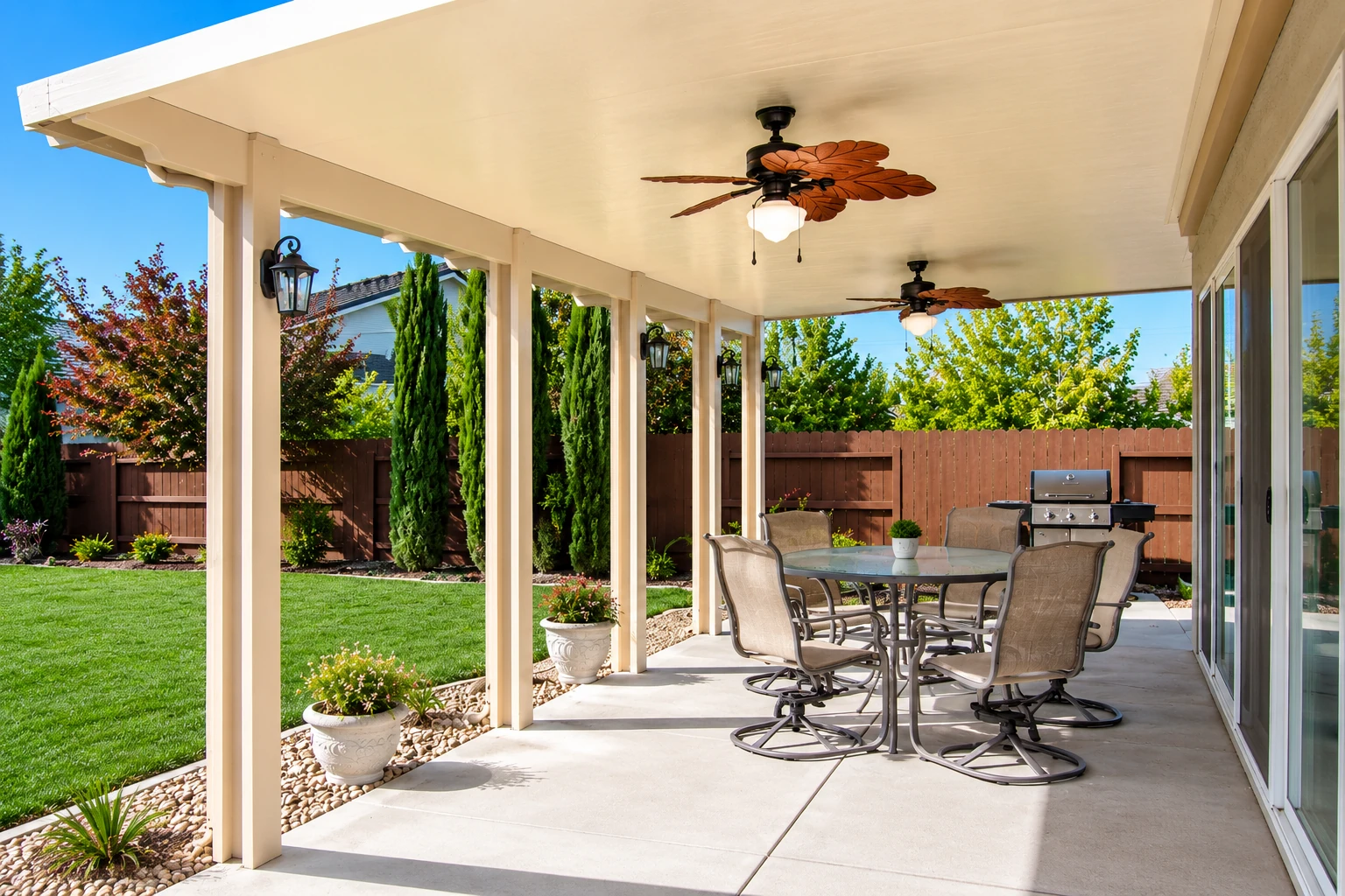 Covered patio with a white roof, ceiling fans and dining set beside a green lawn and fenced garden.