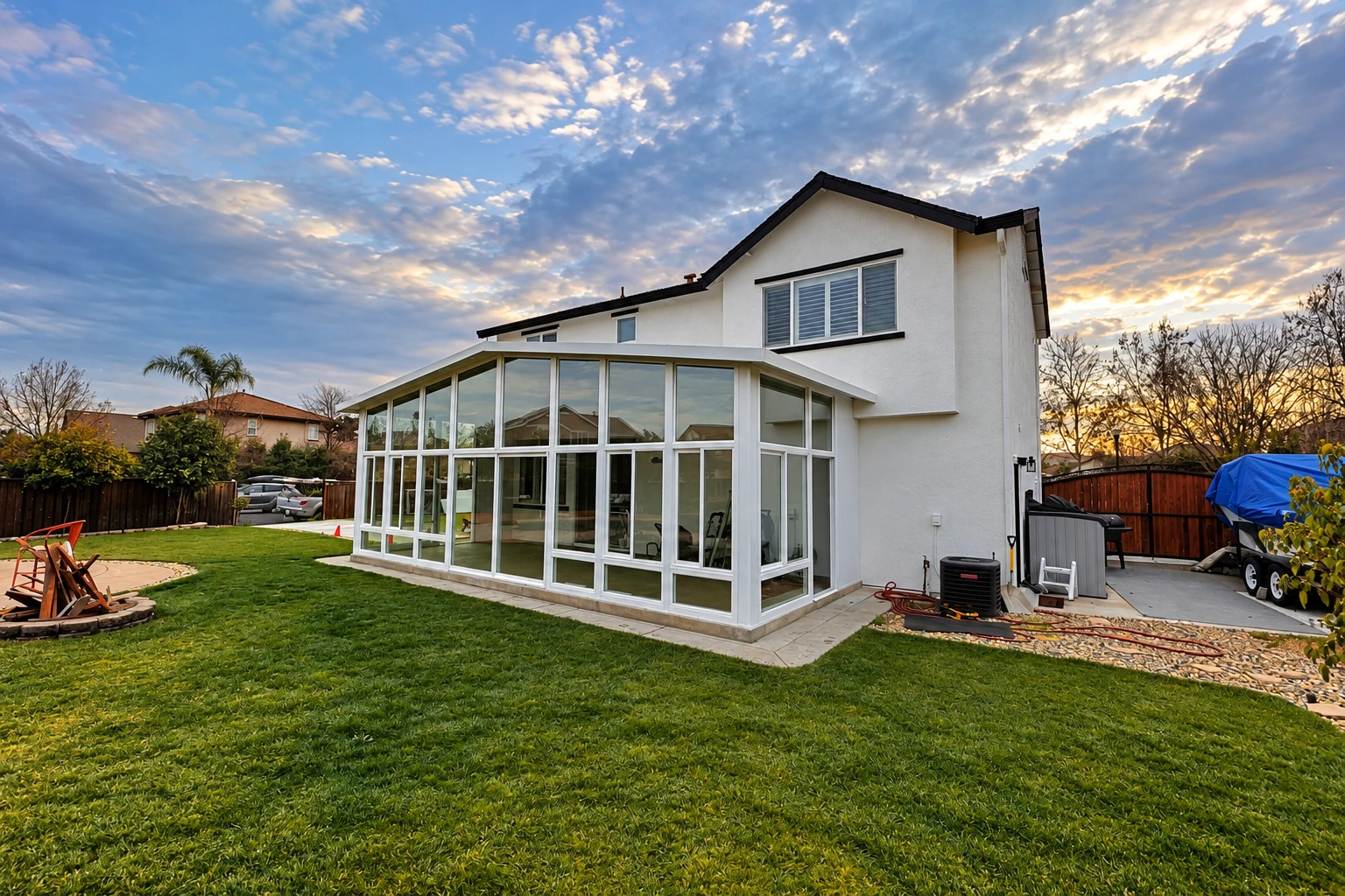 Glass sunroom addition with white framing extends from a two-story home into a broad lawn at sunset.