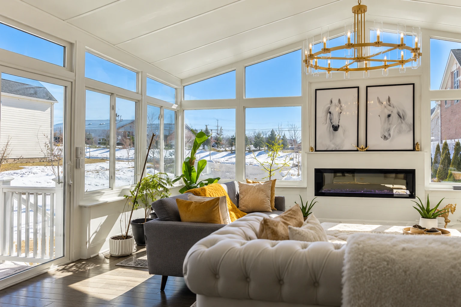 Bright sunroom with white-framed windows, gray seating, a fireplace and snowy views outside.