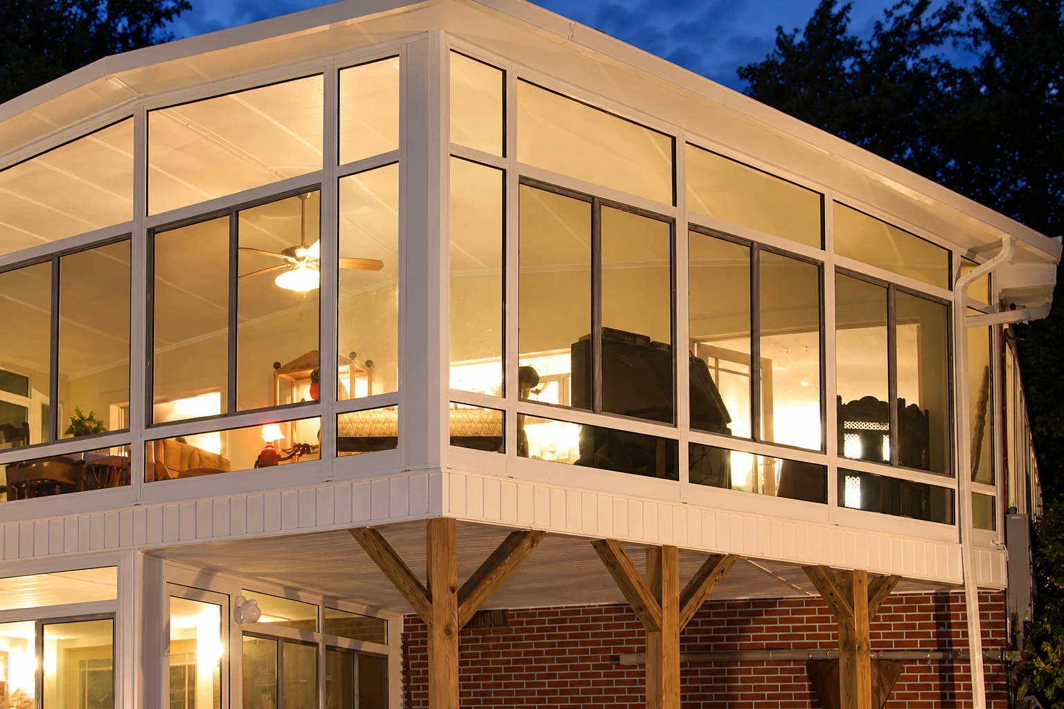 A brightly lit, glass-enclosed sunroom extends from a raised house at dusk, with white framing and a gabled roof.