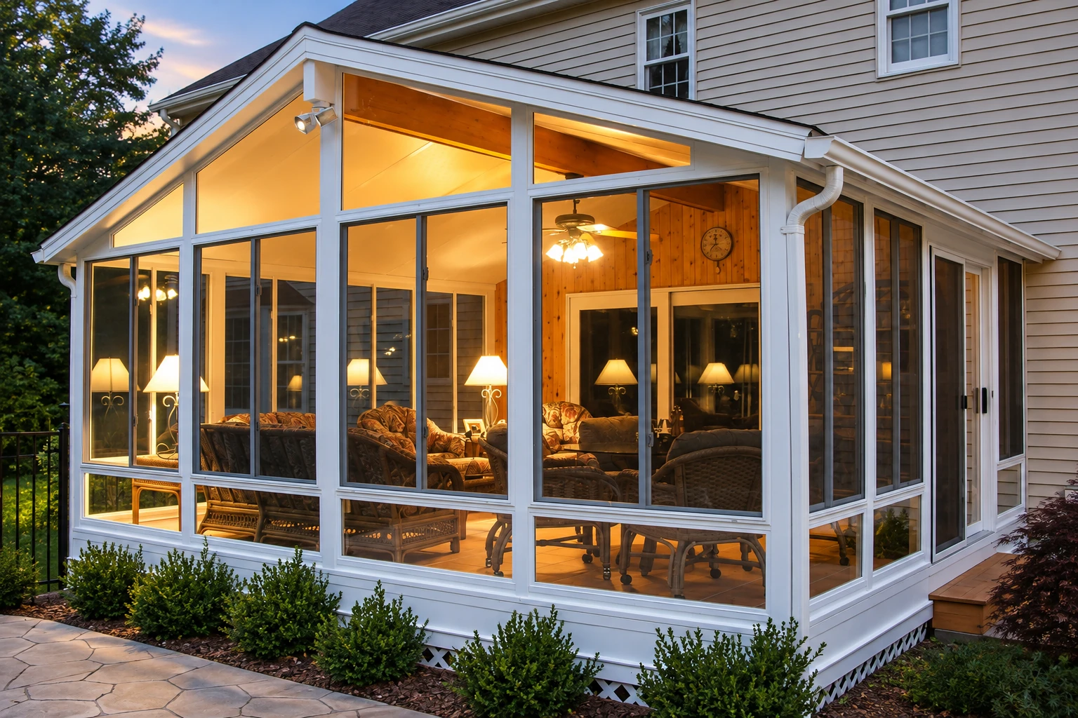 A white-framed sunroom with a peaked roof glows warmly beside a beige-sided house at dusk.