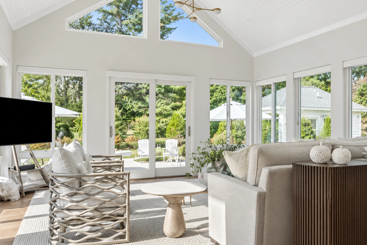 A bright sunroom interior with white walls, vaulted ceiling, large windows, and neutral seating overlooking a garden.